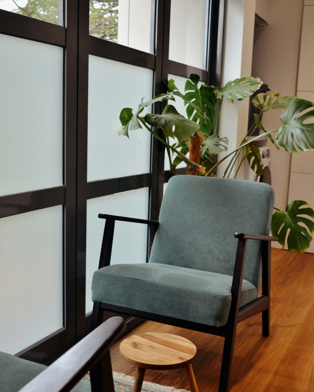 A calm, sunlit therapy room with a soft sage-green armchair, a small wooden side table, and a leafy plant by the window